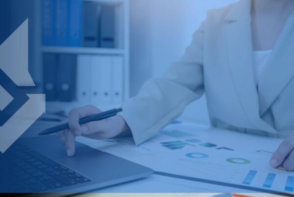 A person in a beige suit reviews charts and graphs on paper, using a laptop, in an office with binders on shelves. Professional and analytical.