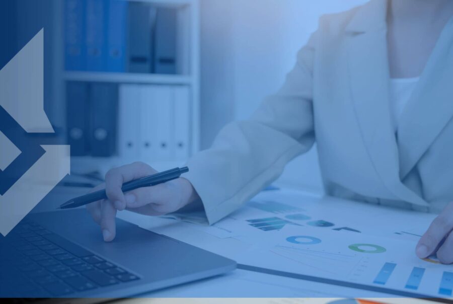 A person in a beige suit reviews charts and graphs on paper, using a laptop, in an office with binders on shelves. Professional and analytical.