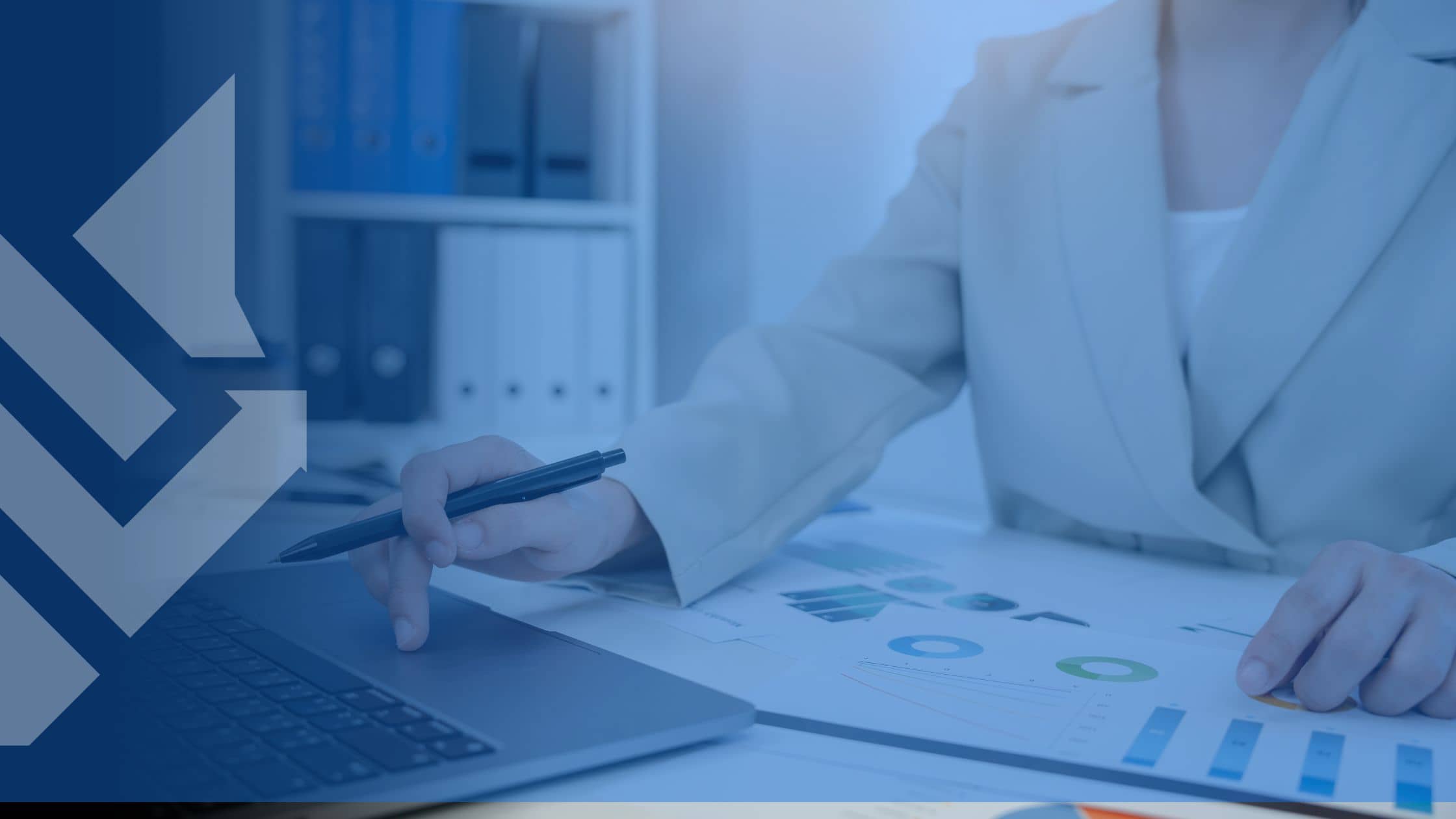 A person in a beige suit reviews charts and graphs on paper, using a laptop, in an office with binders on shelves. Professional and analytical.