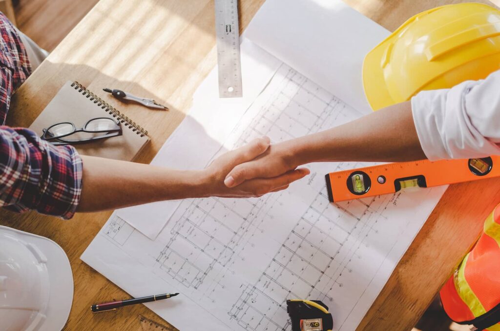 Two contractors shaking hands over blueprints and tools