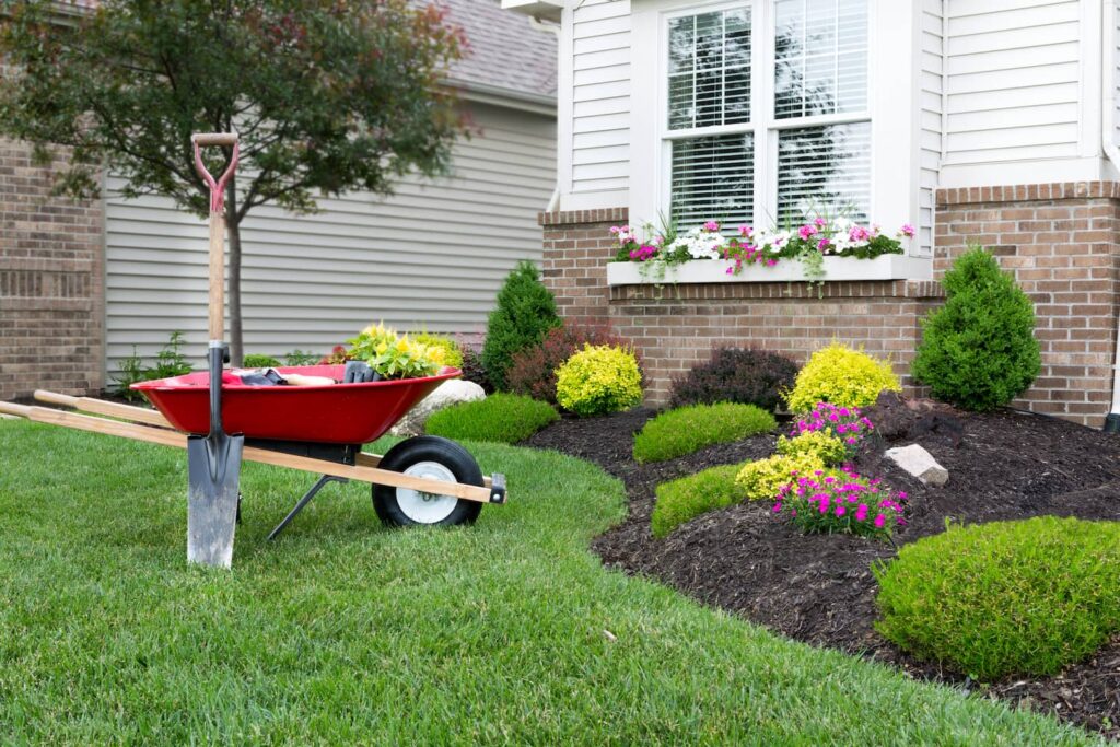 Landscaping project in progress with a red wheelbarrow, shovel, and garden tools on a manicured lawn in front of a home—ideal for a digital marketing agency for landscapers.