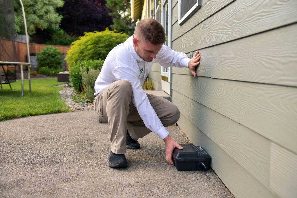 Natura Pest Control technician checking a rodent bait station along the side of a home, Vancouver, WA